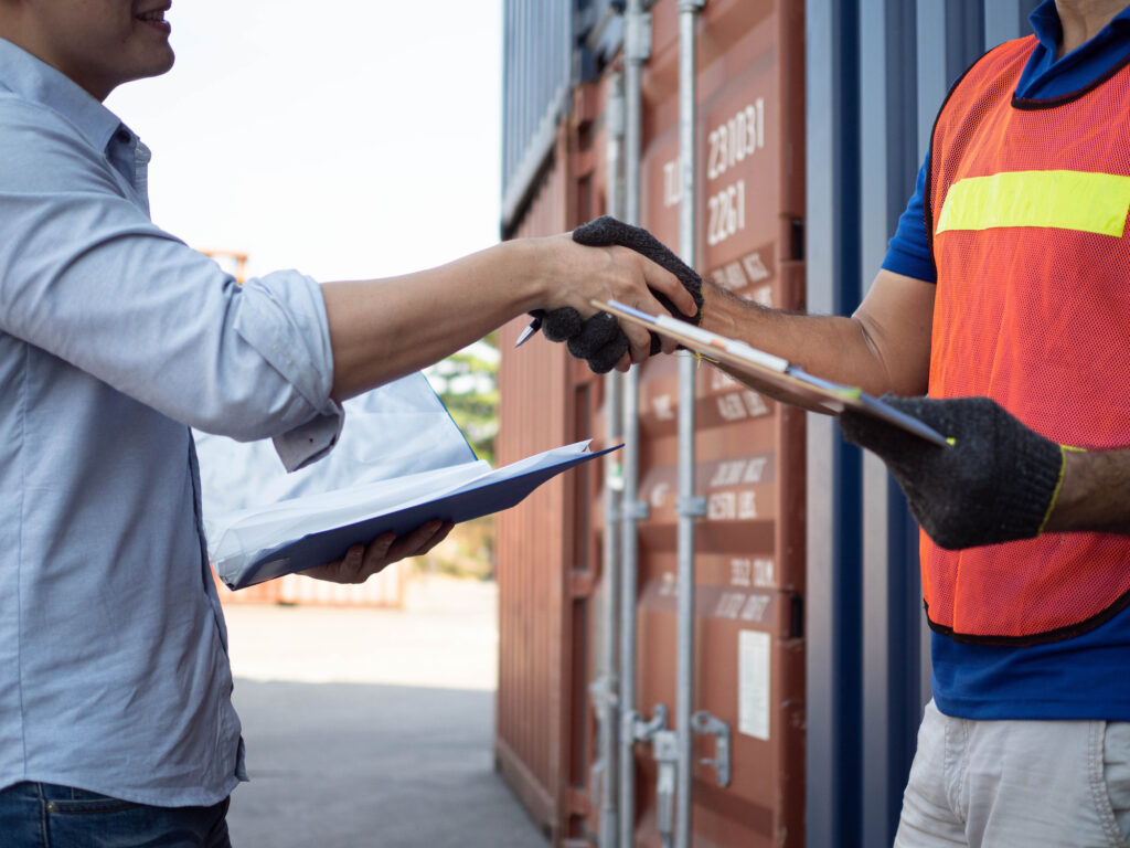 A customs broker shaking hands with a worker in a shipping container yard.